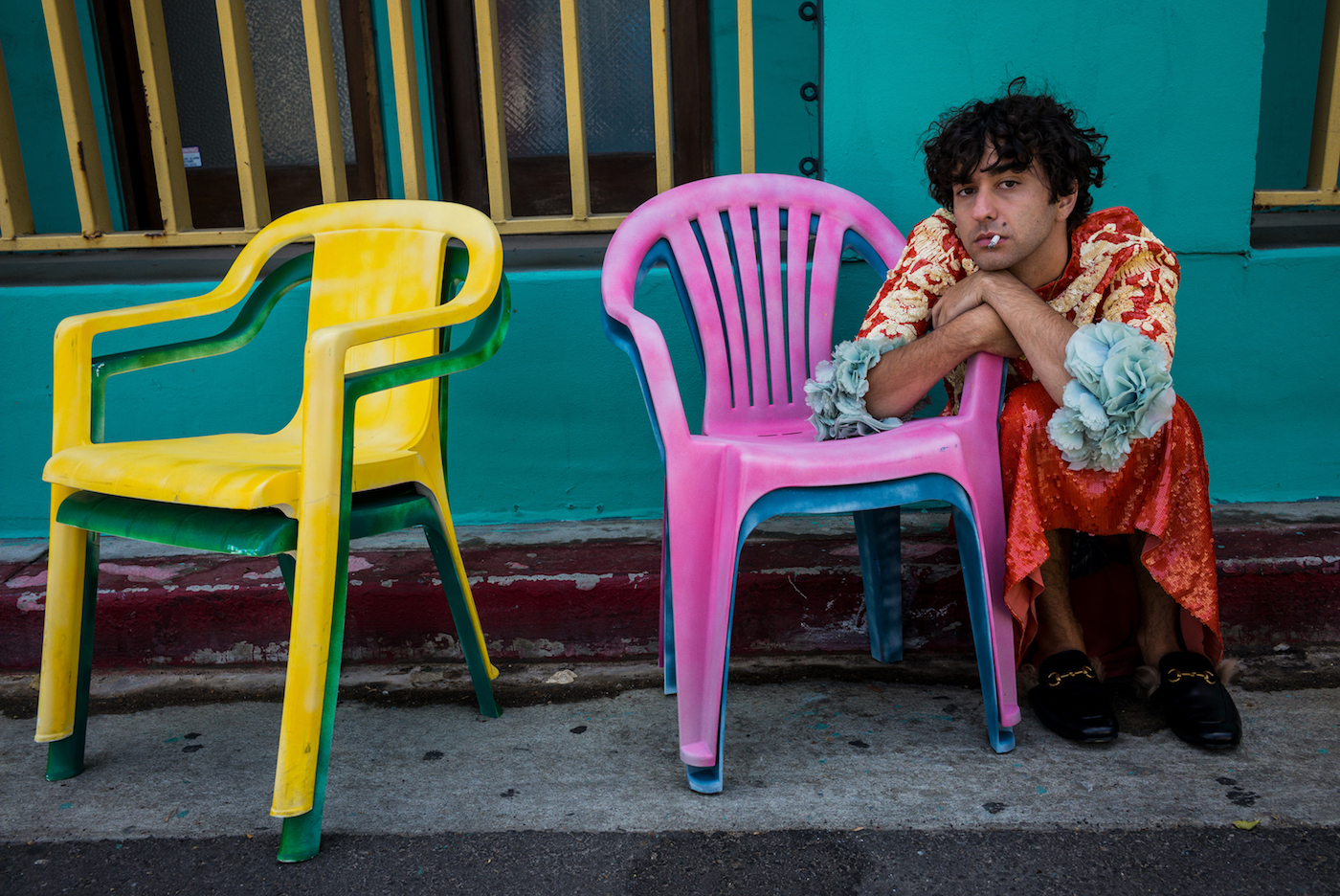 Man in dress, with cigarette, crouching next to plastic chairs