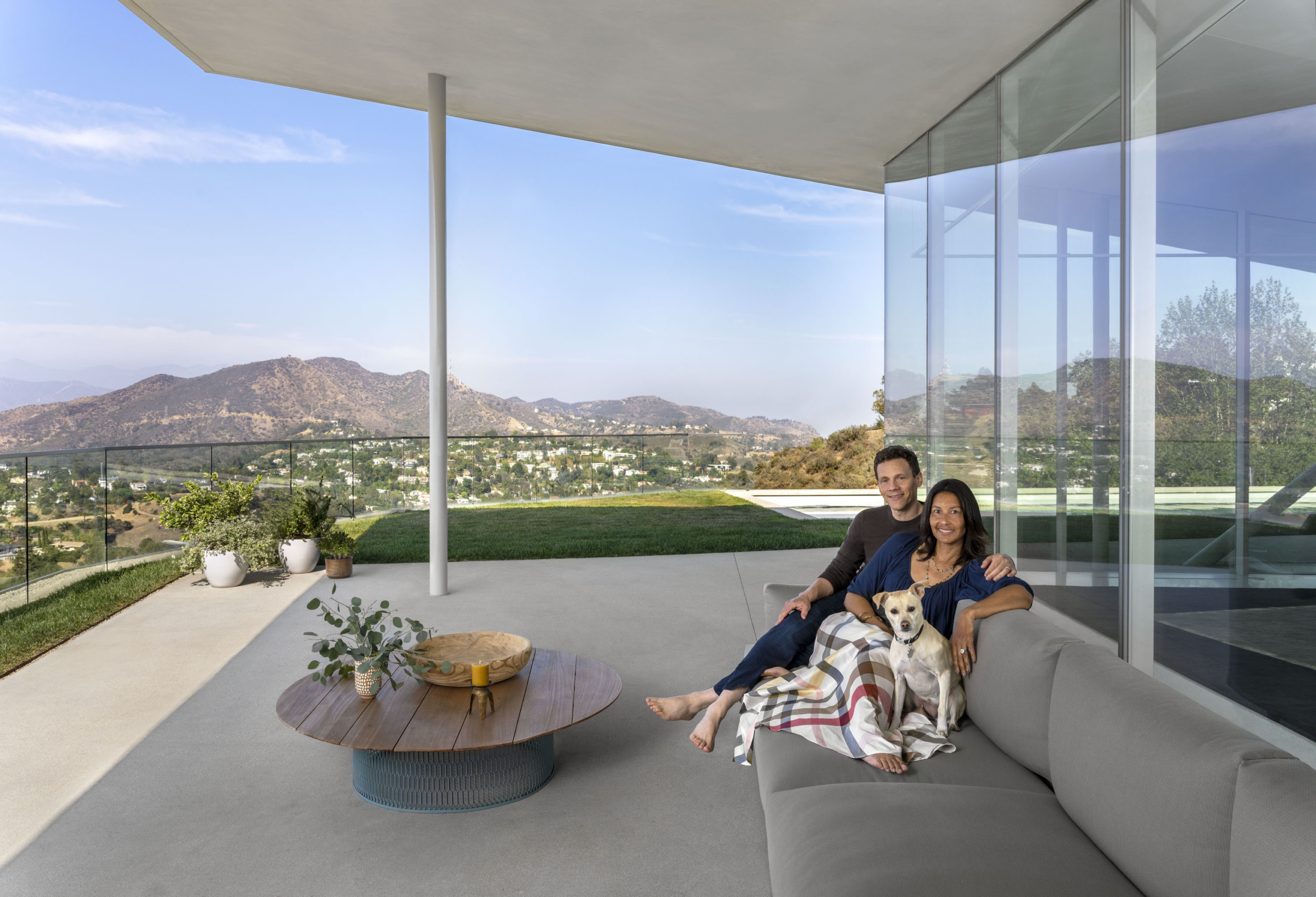 smiling couple sitting on outdoor couch on balcony of modern home, with mountains of LA in background