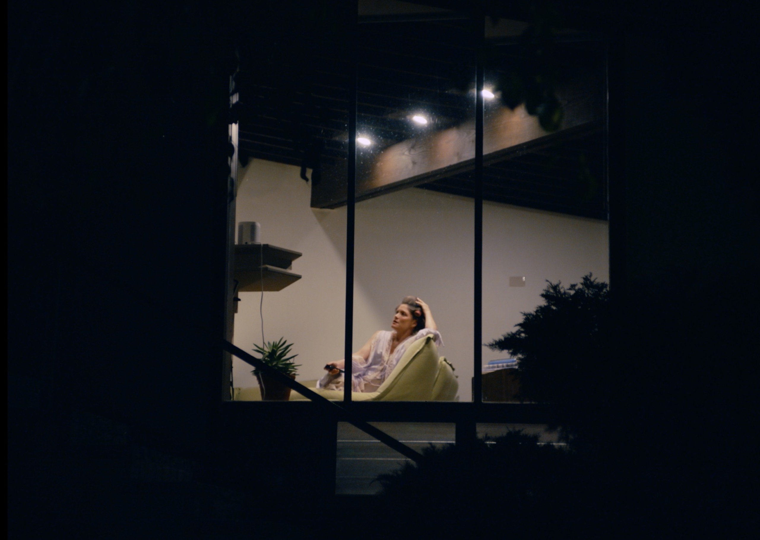 shot of woman from outside her home sitting on a chair with a drink at night