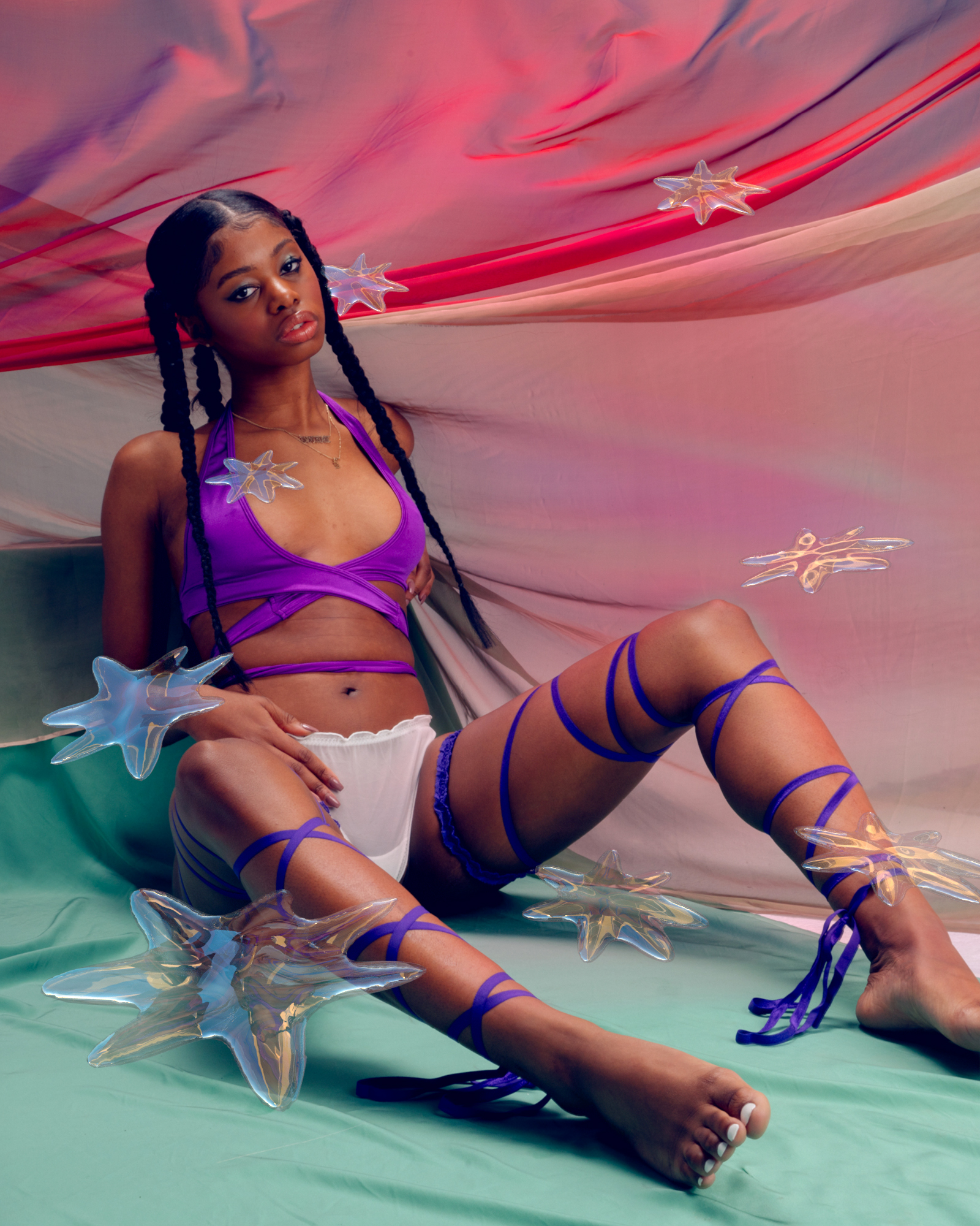 model wearing siren basics white thong and other purple accessories and garments sits in colorful set with stars transposed onto theimage