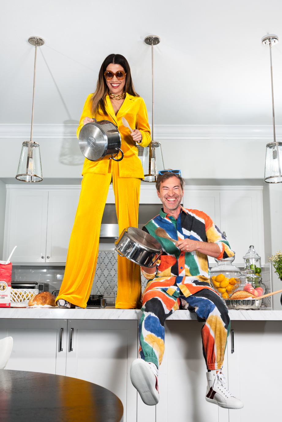 Mark Duplass sits and Natalie Morales stands on a kitchen countertop holding pots and laughing.