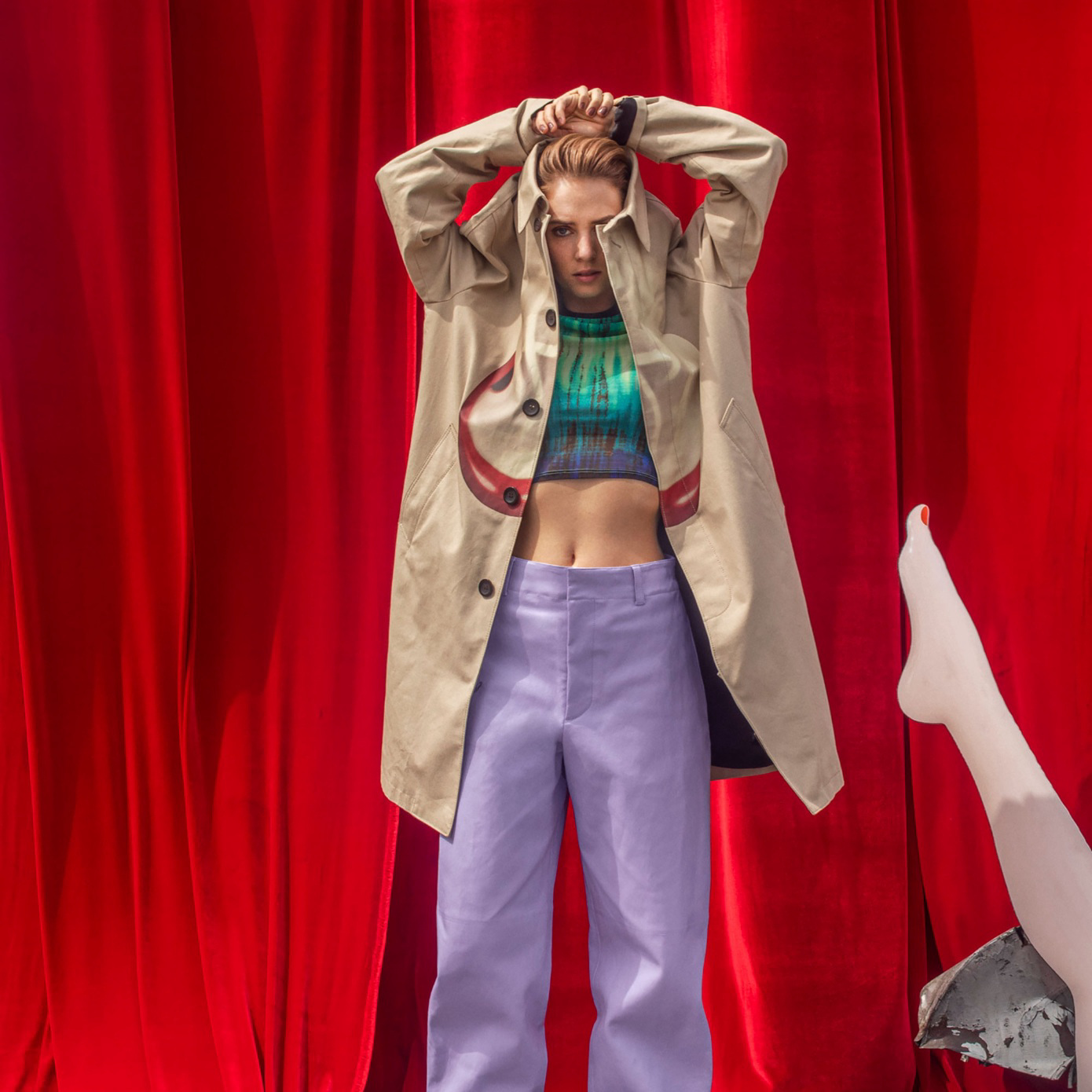 woman in coat poses with hands behind her head in front of red curtains