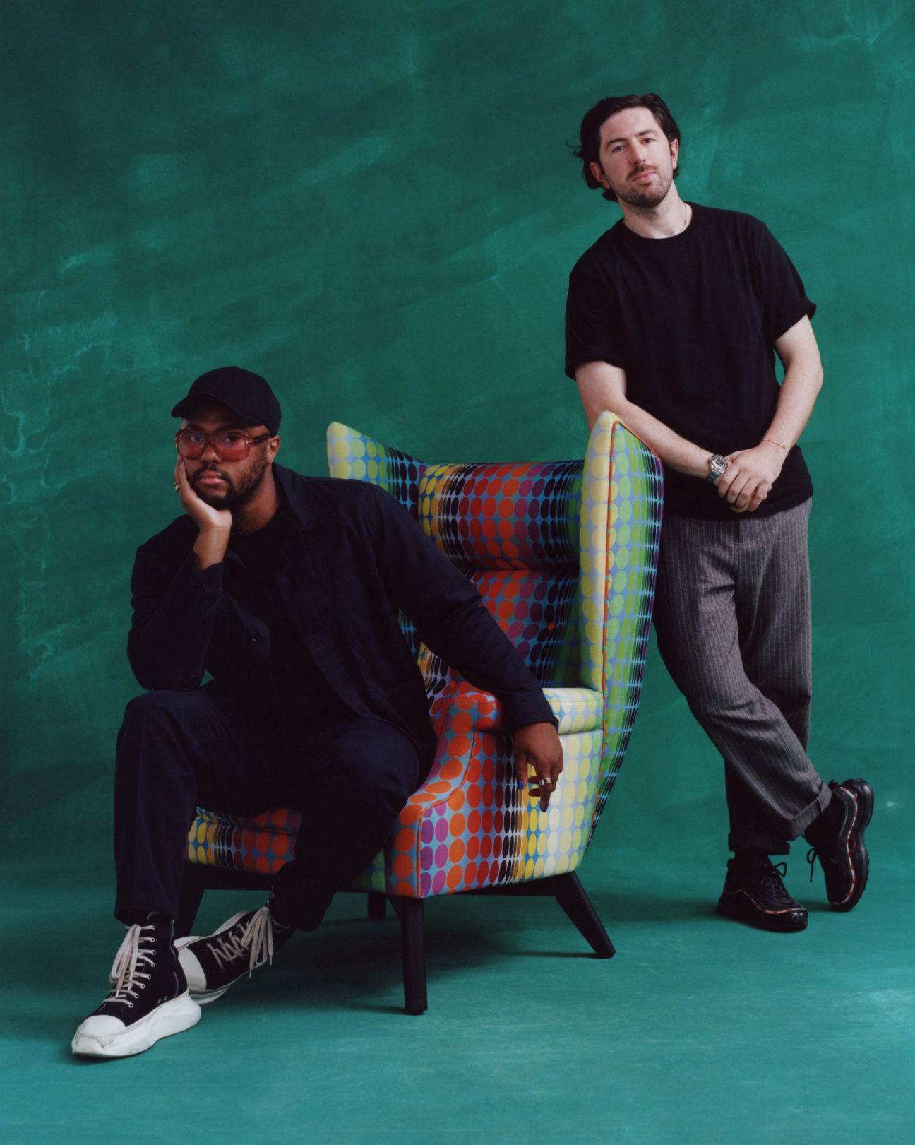 Christopher John Rogers and Ciarán McGuigan photographed with multicolored circular patterned chair, against a dark green backdrop.