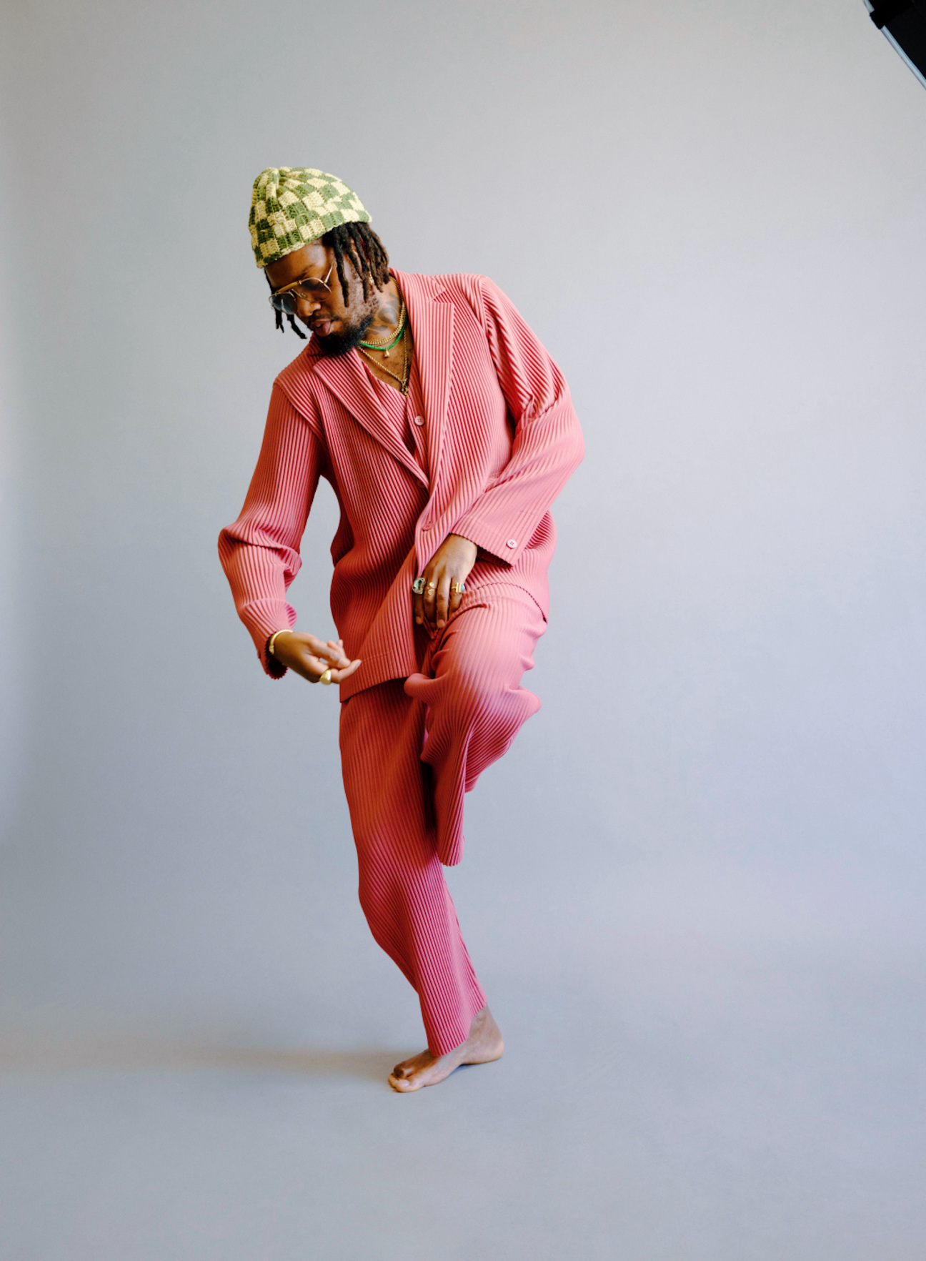 man dances in front of blank background wearing pleated suit and checkered beanie