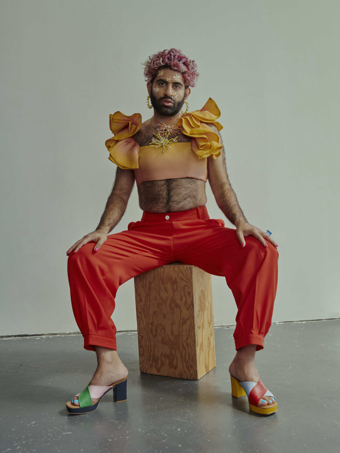 alok vaid menon poses sitting on wood block with legs apart and hands on legs