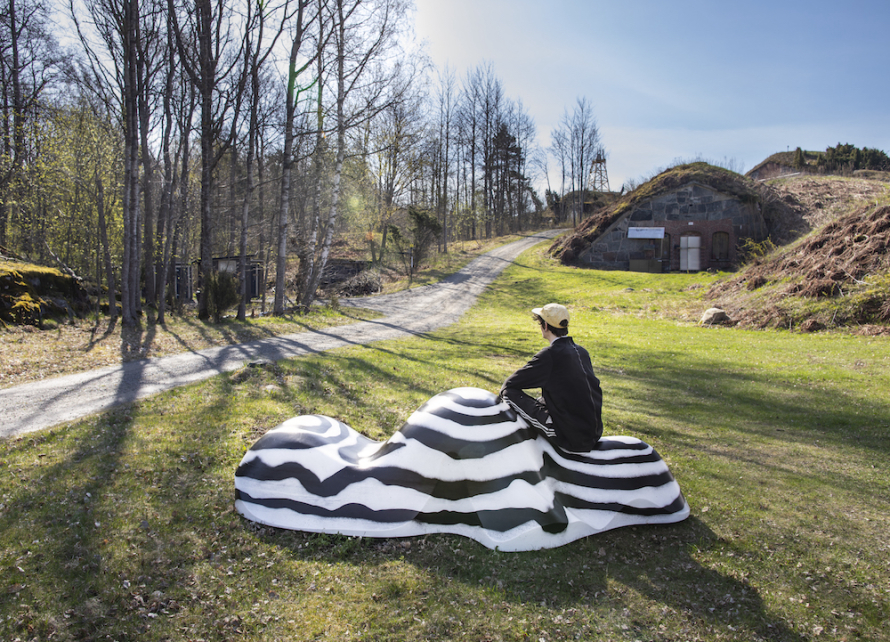 man sits upon striped rock sculpture in wide green hill space