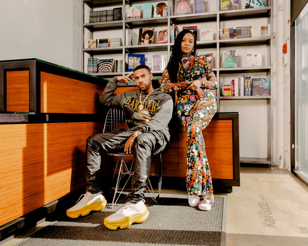 Man sits in chair in front of a bookshelf, woman stands beside him